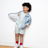 Child wearing a light blue jacket over a patterned shirt and shorts, standing against a white wall.