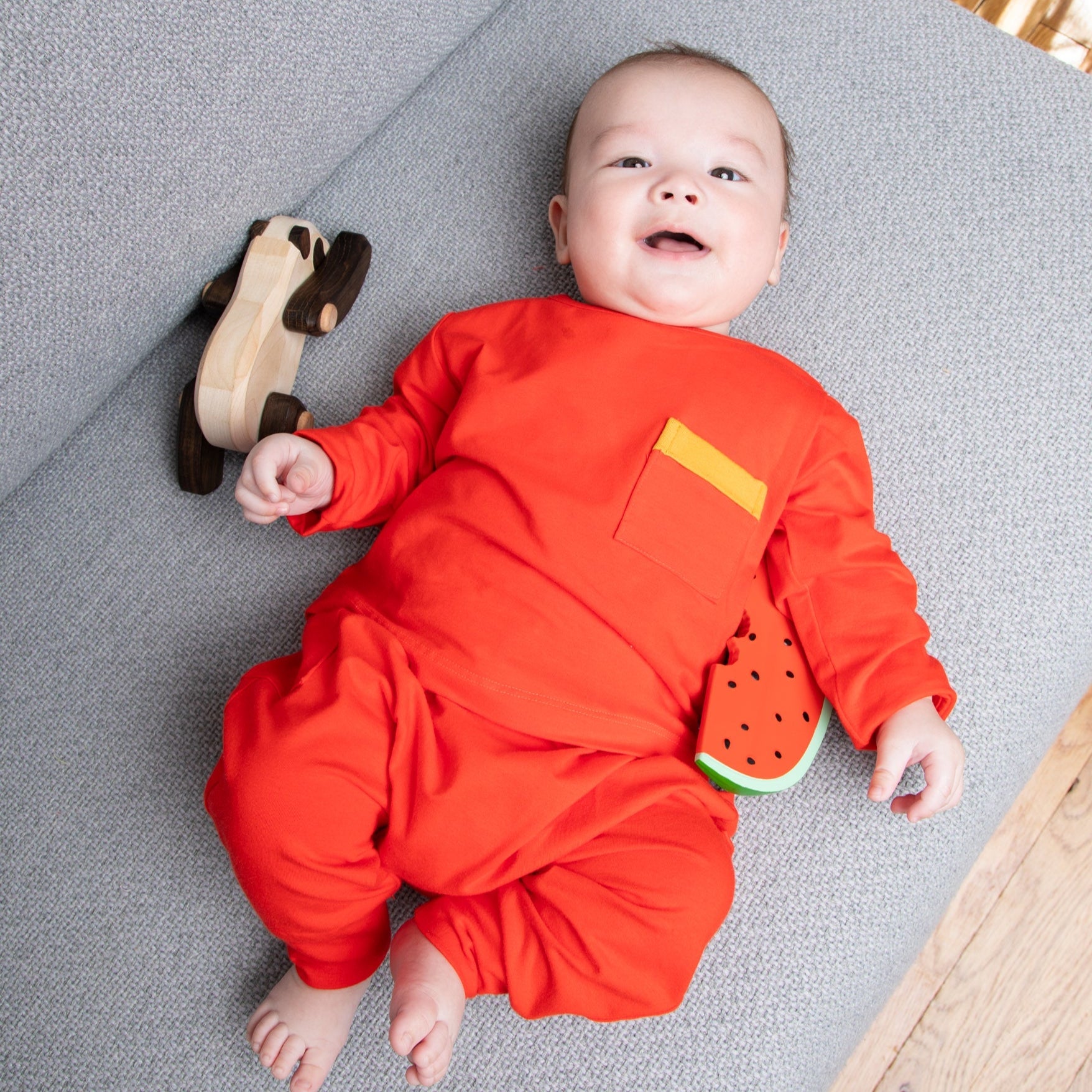 Baby boy lying on a grey couch wearing a vermillion red pants and longsleeve shirt