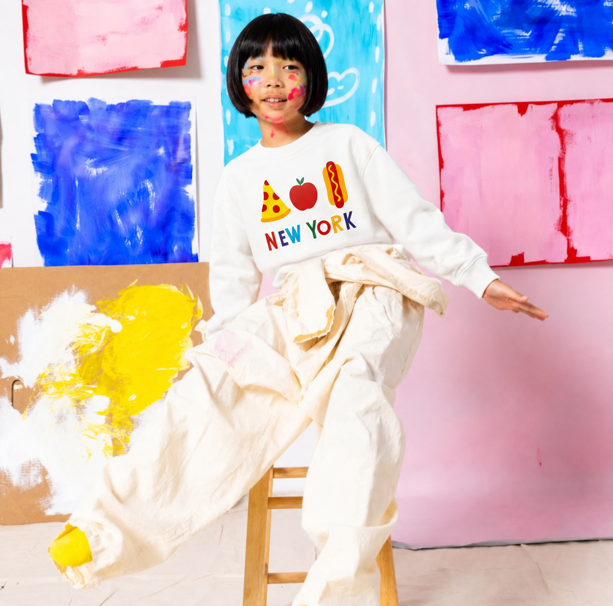A child model wearing a white crewneck sweatshirt with a colorful 'NEW YORK' graphic and matching cream-colored pants, sitting on a wooden stool in front of a pink wall decorated with colorful abstract paintings.