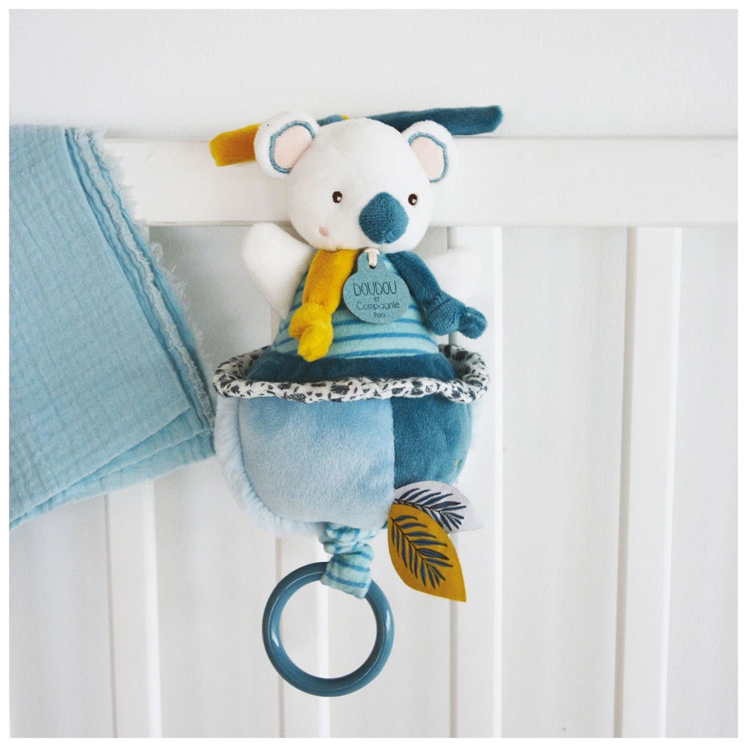 Doudou et Compagnie Yoka the Koala Musical Pull Toy – soft plush koala in teal and white with yellow pull cord and ring, shown with botanical gift box string on a crib.