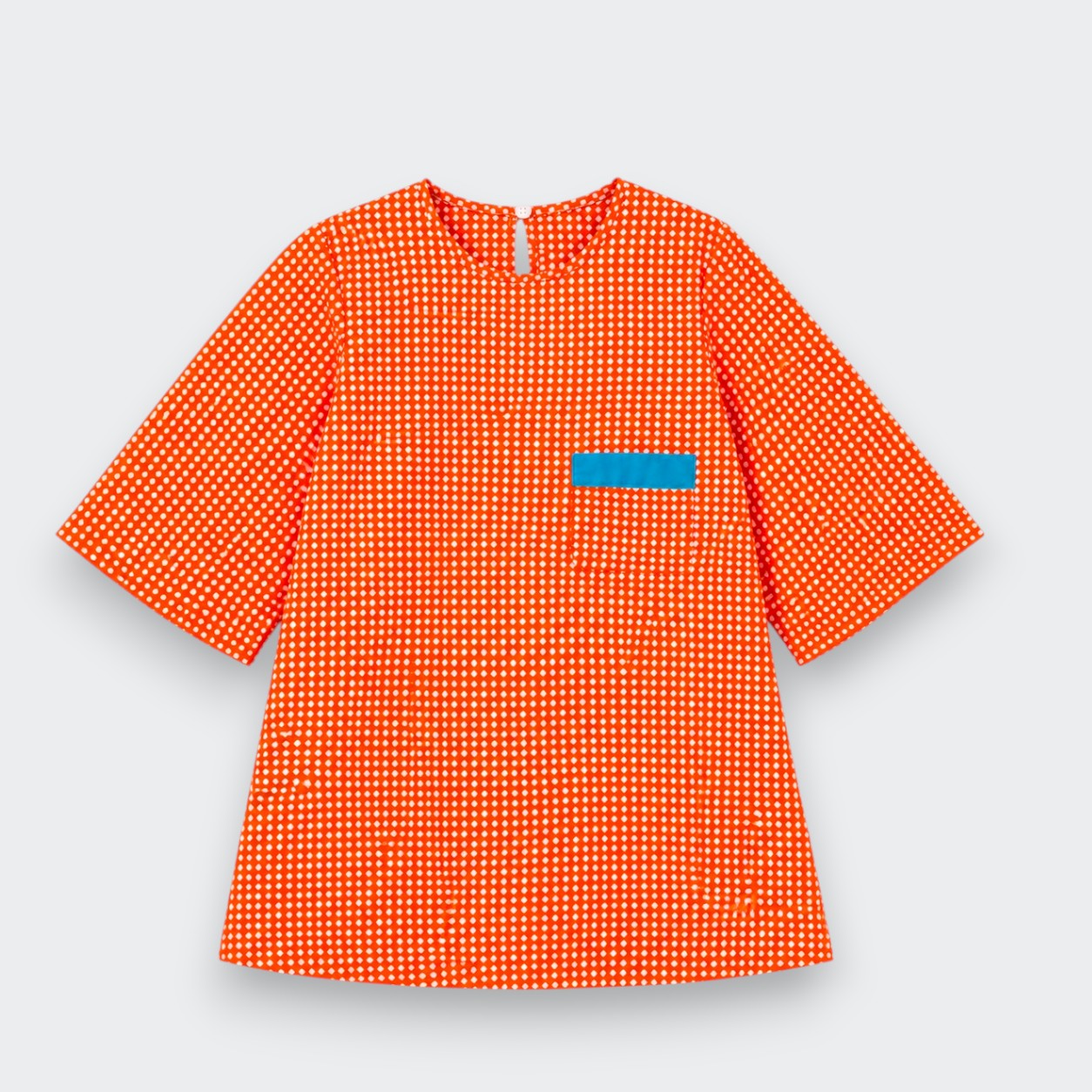 An orange and white polka dot tunic dress with three-quarter sleeves and a small blue rectangular patch on the chest, shown against a light gray background.