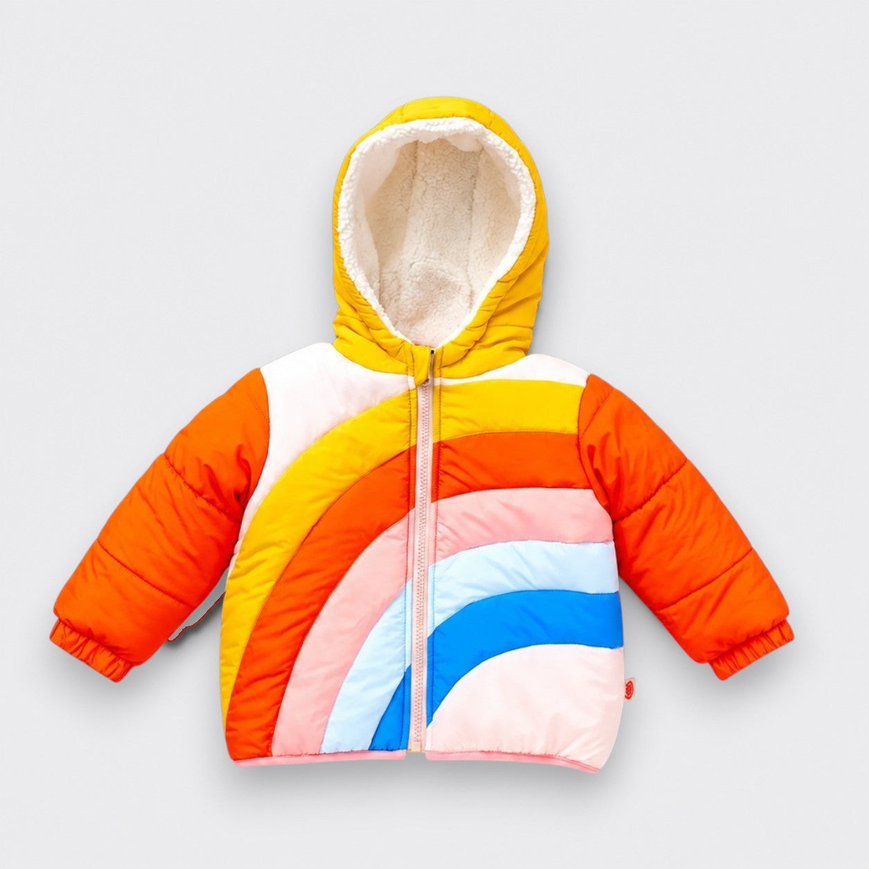 A children’s hooded puffer jacket laid flat on a light gray background. It has a yellow hood with white plush lining, orange quilted sleeves, a pale pink front zipper, and curved rainbow panels in yellow, orange, pink, light blue, and bright blue.