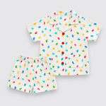 A Dinosaur set consisting of a short-sleeve button-up shirt and matching shorts in an off-white linen-like fabric, decorated with a colorful print of small handprints in blue, green, yellow, and pink. The shirt features red buttons.