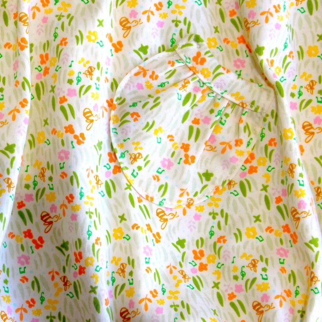 A close-up of a white fabric with a colorful all-over print featuring small flowers, leaves, and whimsical orange motifs, including a heart-shaped patch pocket.