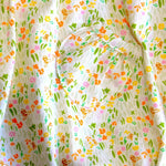 A close-up of a white fabric with a colorful all-over print featuring small flowers, leaves, and whimsical orange motifs, including a heart-shaped patch pocket.