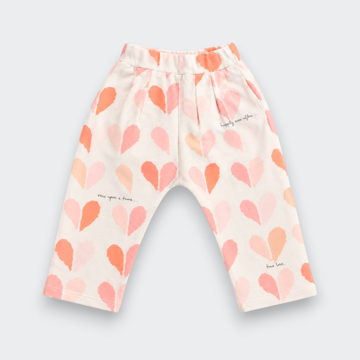 A pair of toddler's elastic-waist pants in a warm off-white fabric, decorated with a pattern of hand-drawn hearts in shades of pink, peach, and orange, with small black script text details.