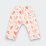 A pair of toddler's elastic-waist pants in a warm off-white fabric, decorated with a pattern of hand-drawn hearts in shades of pink, peach, and orange, with small black script text details.