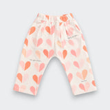 A pair of toddler's elastic-waist pants in a warm off-white fabric, decorated with a pattern of hand-drawn hearts in shades of pink, peach, and orange, with small black script text details.