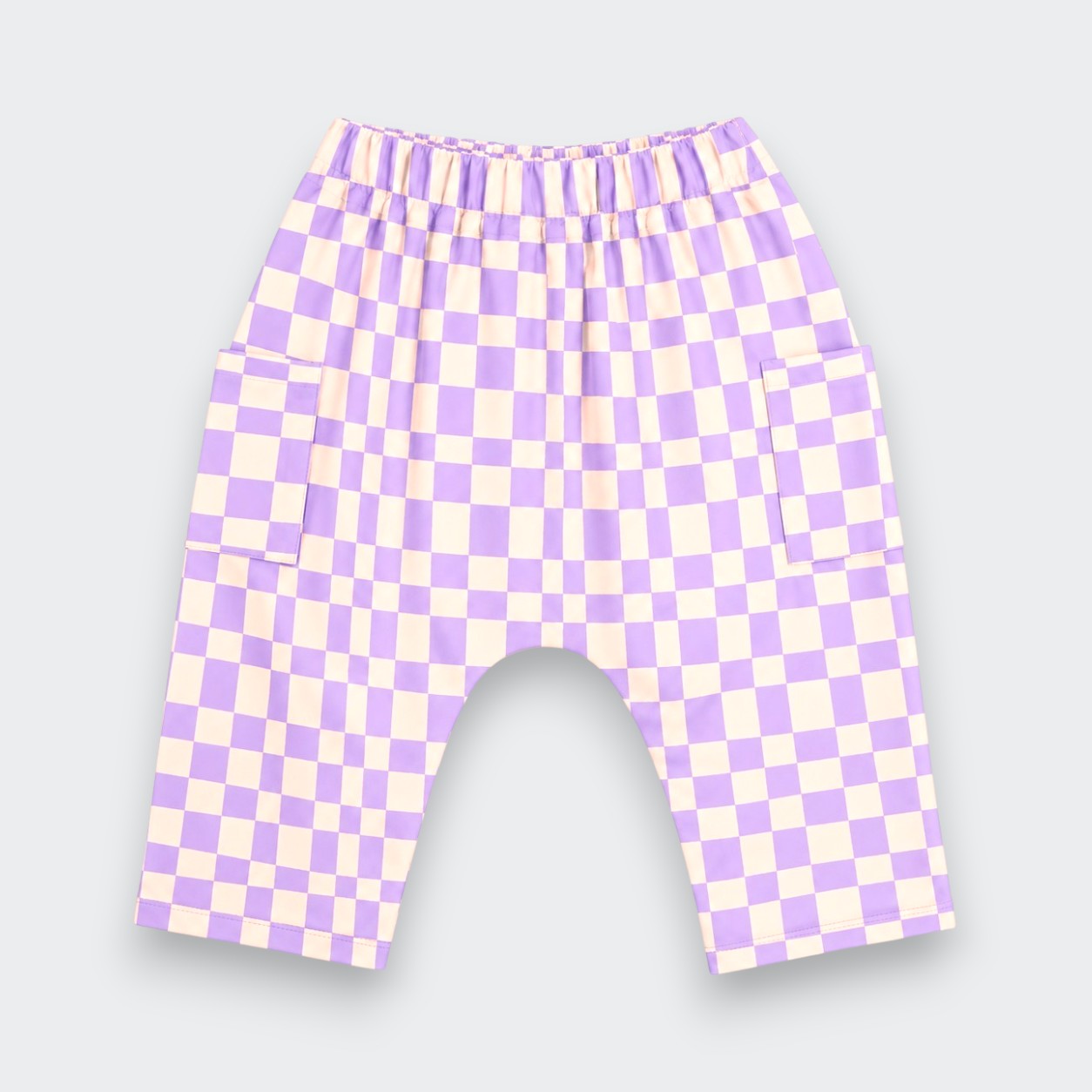A pair of children's shorts featuring a lavender and cream checkerboard pattern, an elastic waistband, and side pockets, shown against a light gray background.