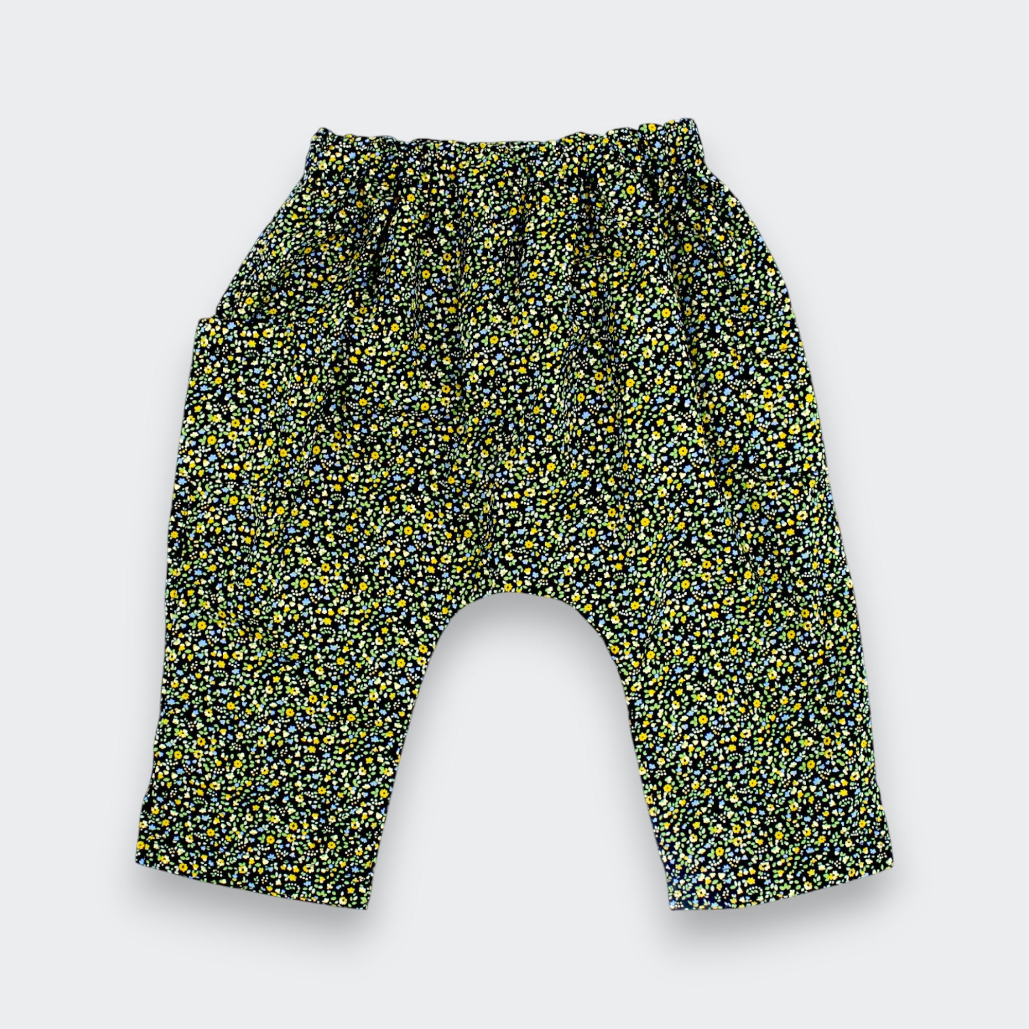 A pair of toddler's harem-style pants with an elastic waistband and a dense yellow and green floral print on a black background, shown flat on a light gray background.