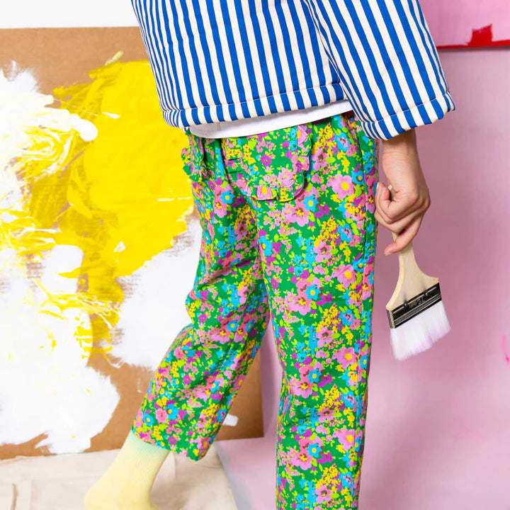 A child wearing bright green floral pants and a blue-and-white striped shirt, holding a paintbrush in a pink studio setting with yellow and white paint strokes in the background.