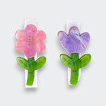 Two flower hair clips on a light gray background: a pink five-petal flower with an orange center and a purple tulip. Each has a green stem and leaves mounted on a white clip.