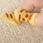 Close-up of a baby wearing yellow socks with rust-brown giraffe-like spots while resting on a cream textured blanket. White grip dots are visible on the sole of one sock.