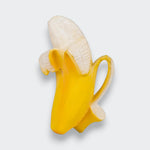 A banana-shaped baby teether on a light gray background. It has a textured cream-colored banana, a bright yellow peel folded back around it, and a loop handle on one side.