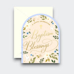An arch-shaped baptism greeting card reading “Baptism Blessings” in gold script, with a small cross, delicate flowers, green leaves, and a pale blue border. A cream envelope sits behind it.