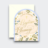 An arch-shaped baptism greeting card reading “Baptism Blessings” in gold script, with a small cross, delicate flowers, green leaves, and a pale blue border. A cream envelope sits behind it.
