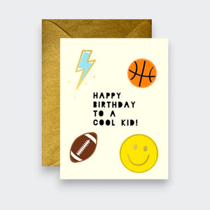 A cream birthday card reading “Happy Birthday to a Cool Kid!” with drawings of a blue lightning bolt, basketball, football, and yellow smiley face. A kraft envelope sits behind it.