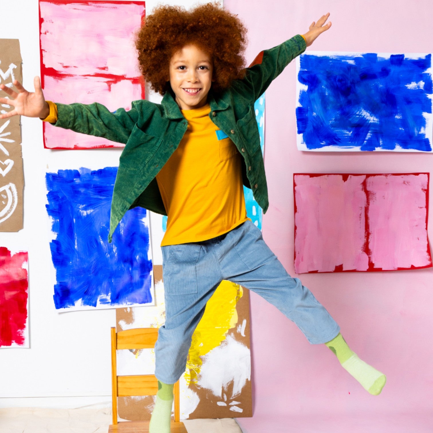Child wearing Little Moony corduroy blue pants jumping in front of colorful abstract paintings on a pink wall