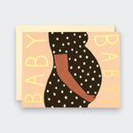 A peach greeting card with a stylized illustration of a pregnant person in a black polka-dot dress, hands resting on their belly. “BABY!” runs vertically along both sides, with a cream envelope behind the card.