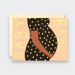 A peach greeting card with a stylized illustration of a pregnant person in a black polka-dot dress, hands resting on their belly. “BABY!” runs vertically along both sides, with a cream envelope behind the card.