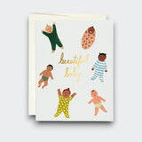 A white greeting card reading “beautiful baby” in gold script, surrounded by illustrations of six babies in colorful pajamas, a swaddle, and patterned diapers. A cream envelope sits behind it.