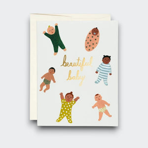 A white greeting card reading “beautiful baby” in gold script, surrounded by illustrations of six babies in colorful pajamas, a swaddle, and patterned diapers. A cream envelope sits behind it.