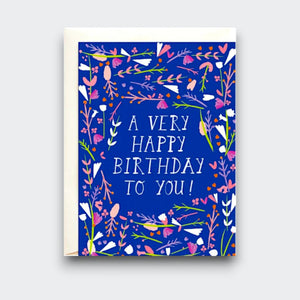A bright blue birthday card reading “A very happy birthday to you!” in white lettering, framed by colorful pink, white, orange, and green flowers and leaves. A cream envelope sits behind it.
