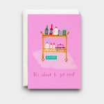 A pink greeting card reading “It’s about to get real,” with a playful illustration of a rolling cart stocked with bottles, baby supplies, and a box labeled “DIAPERS.” A cream envelope sits behind it.