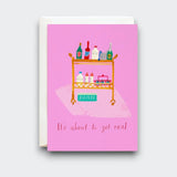 A pink greeting card reading “It’s about to get real,” with a playful illustration of a rolling cart stocked with bottles, baby supplies, and a box labeled “DIAPERS.” A cream envelope sits behind it.