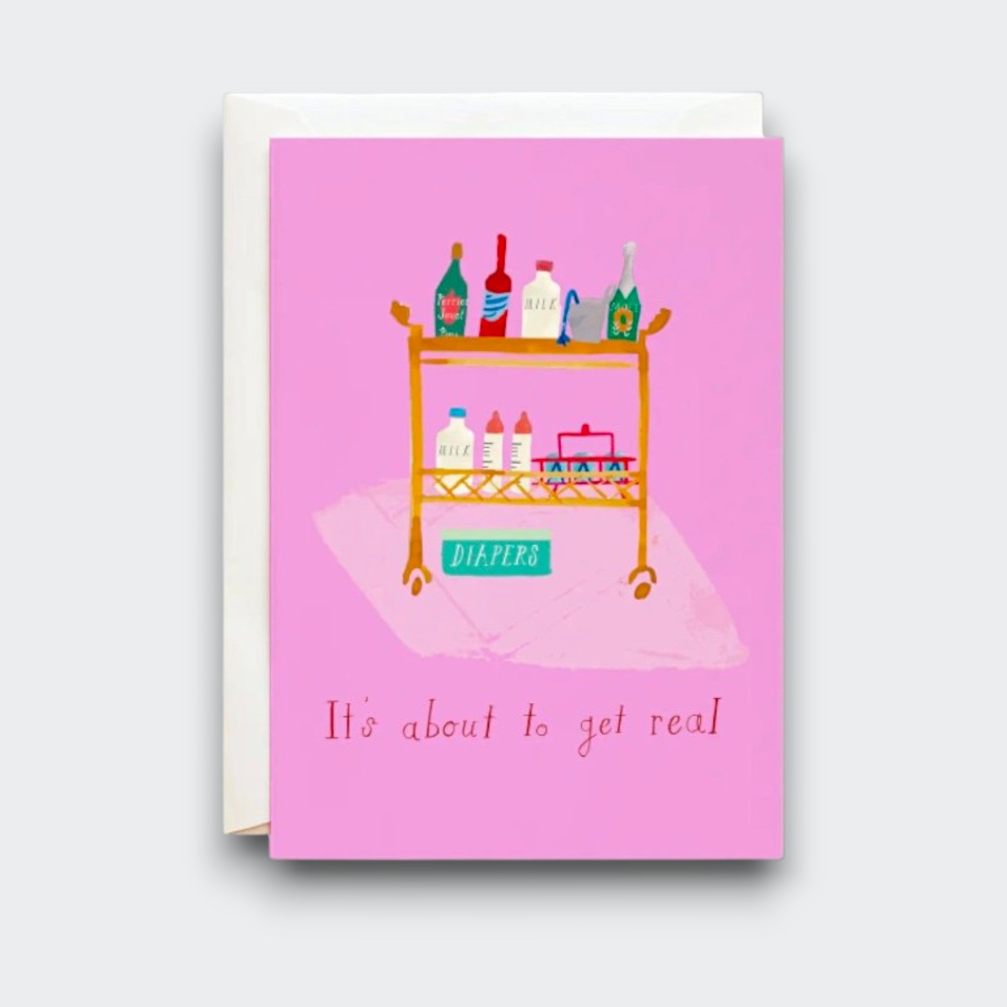 A pink greeting card reading “It’s about to get real,” with a playful illustration of a rolling cart stocked with bottles, baby supplies, and a box labeled “DIAPERS.” A cream envelope sits behind it.