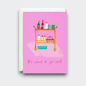 A pink greeting card reading “It’s about to get real,” with a playful illustration of a rolling cart stocked with bottles, baby supplies, and a box labeled “DIAPERS.” A cream envelope sits behind it.