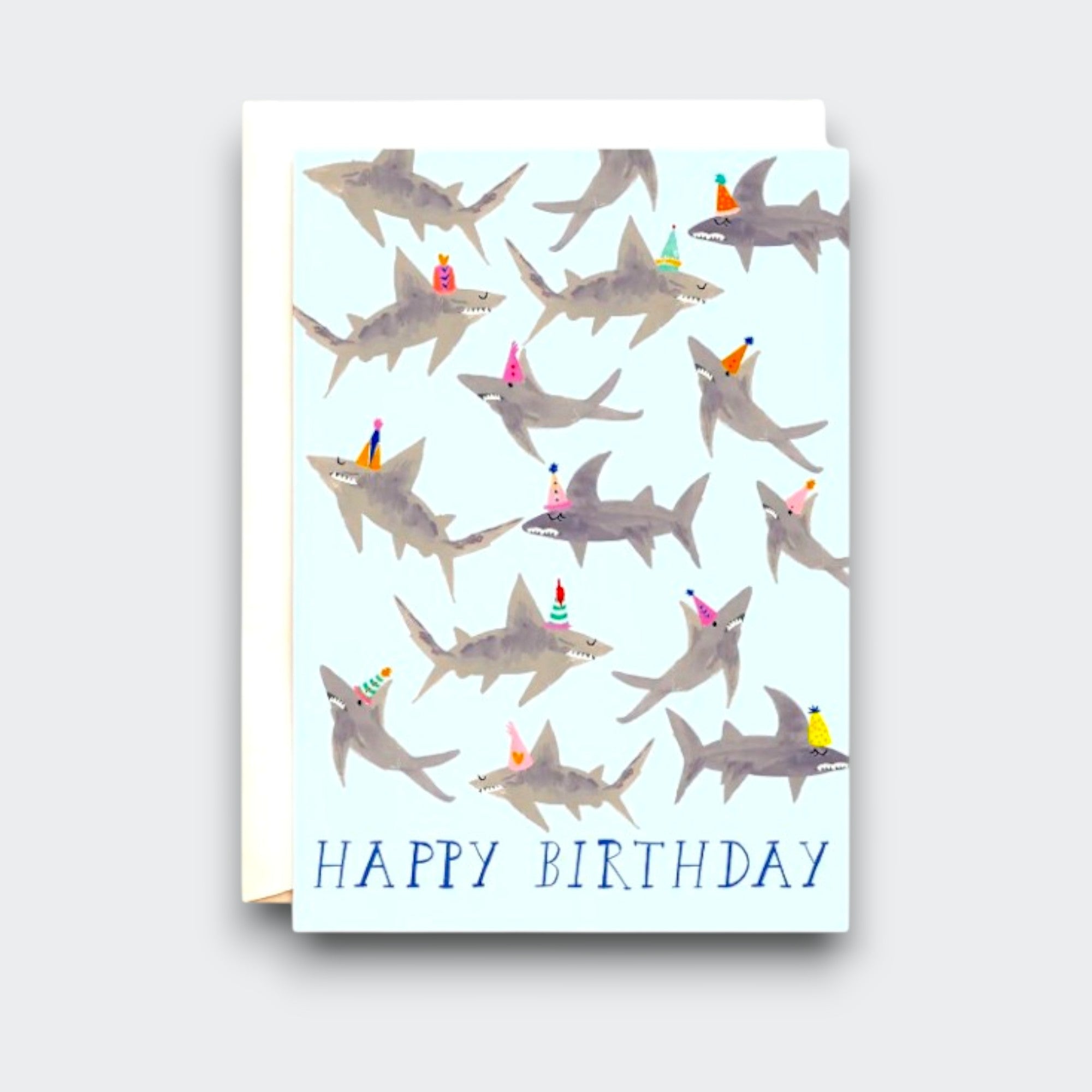 A pale blue birthday card covered with illustrated gray sharks wearing colorful party hats. “HAPPY BIRTHDAY” is printed along the bottom, with a white envelope behind the card.