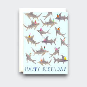 A pale blue birthday card covered with illustrated gray sharks wearing colorful party hats. “HAPPY BIRTHDAY” is printed along the bottom, with a white envelope behind the card.