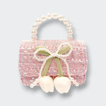 A small pink tweed purse with a rounded pearl-bead handle. A pearl-trimmed bow and two dangling white flower buds decorate the front, photographed against a light gray background.