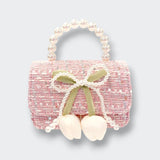 A small pink tweed purse with a rounded pearl-bead handle. A pearl-trimmed bow and two dangling white flower buds decorate the front, photographed against a light gray background.