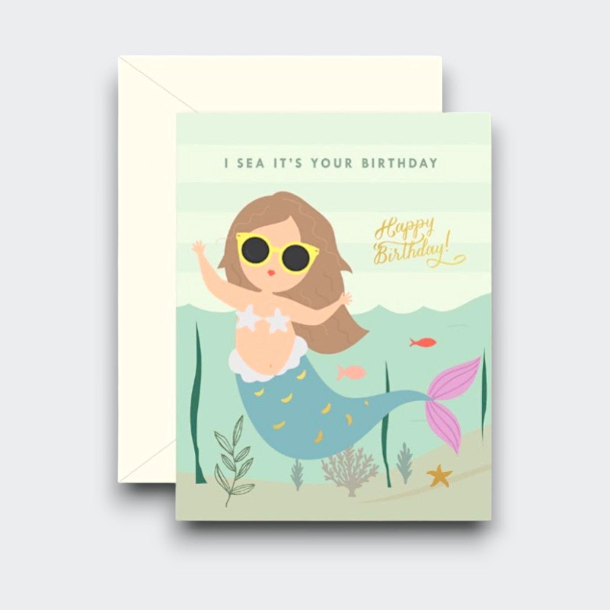 A birthday card showing a mermaid in yellow sunglasses swimming among fish and sea plants. The text reads “I sea it’s your birthday,” with “Happy Birthday!” in gold script. A cream envelope sits behind it.