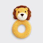 A crocheted lion baby rattle on a light gray background. Its yellow ring handle sits beneath a lion’s face with a brown mane, small round ears, a white muzzle, and a dark embroidered nose.