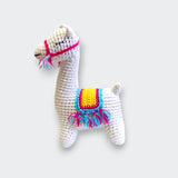 A white crocheted llama rattle shown from the side against a light gray background. It has embroidered closed eyes, a bright pink bridle, and a yellow saddle trimmed in blue and pink with colorful