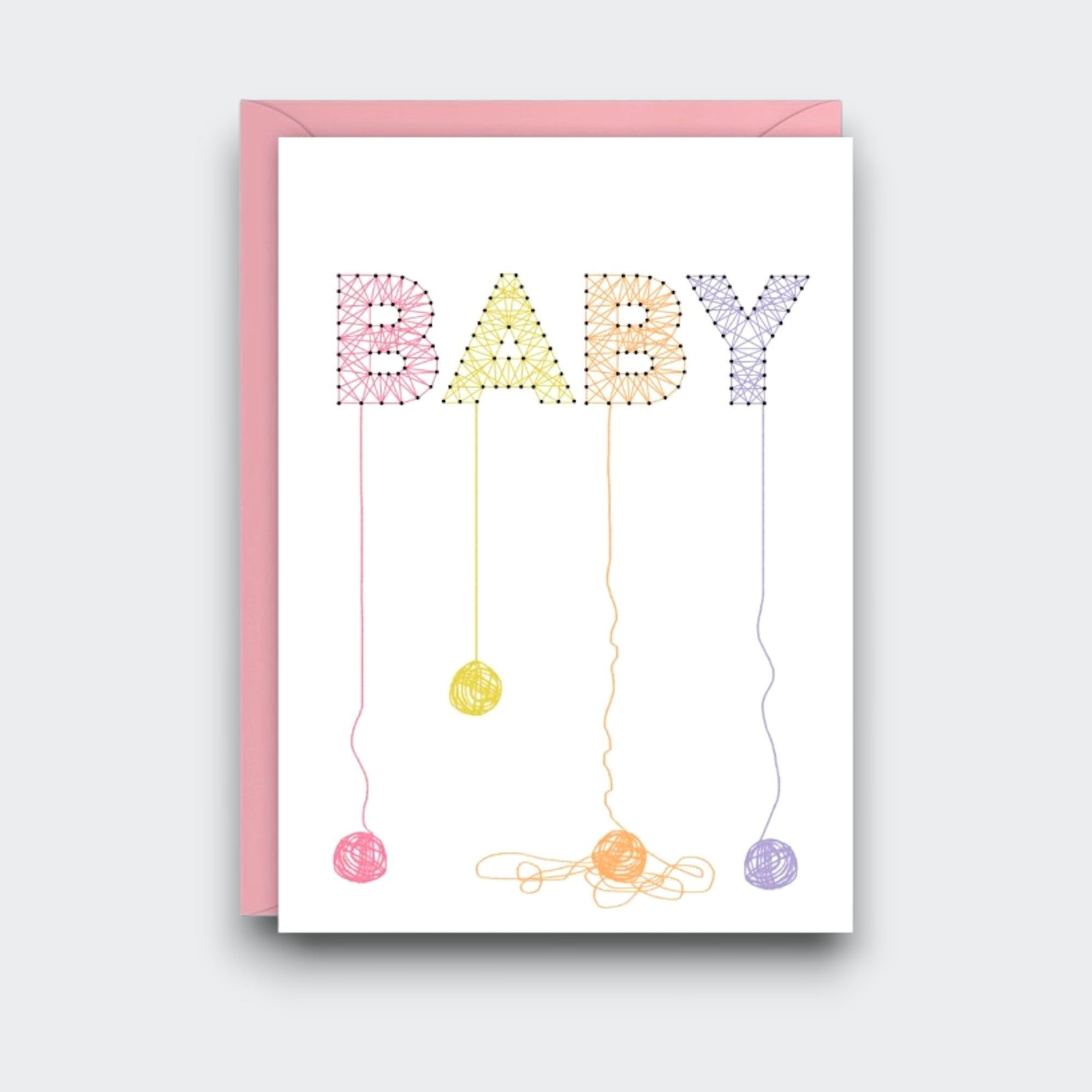 A white baby greeting card with “BABY” designed like pastel string art, each letter trailing a thread and small ball of yarn. A pink envelope sits behind the