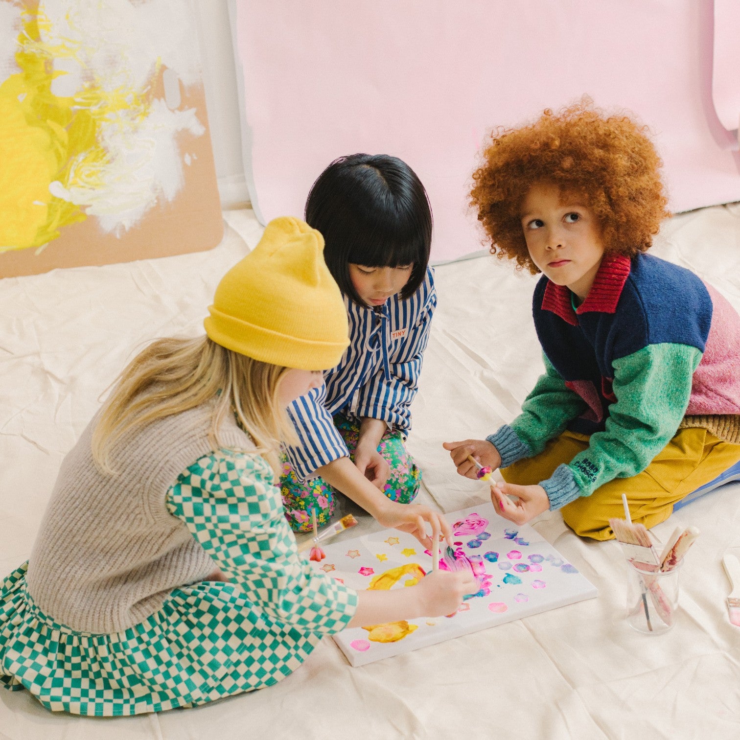 Three children wearing little moony children clothes in new york sitting on a bed playing with colorful toys.
