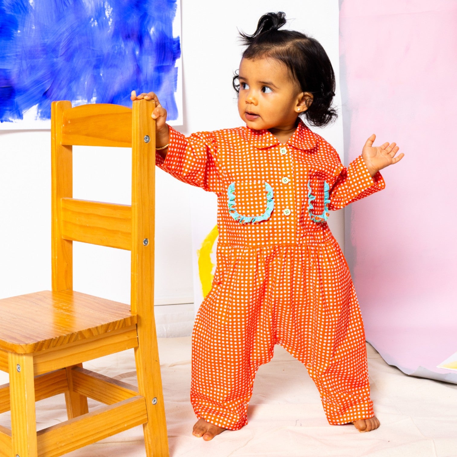 Baby Girl in Little Moony Cherry Dot Cotton Ruffle Pocket Romper beside a chair