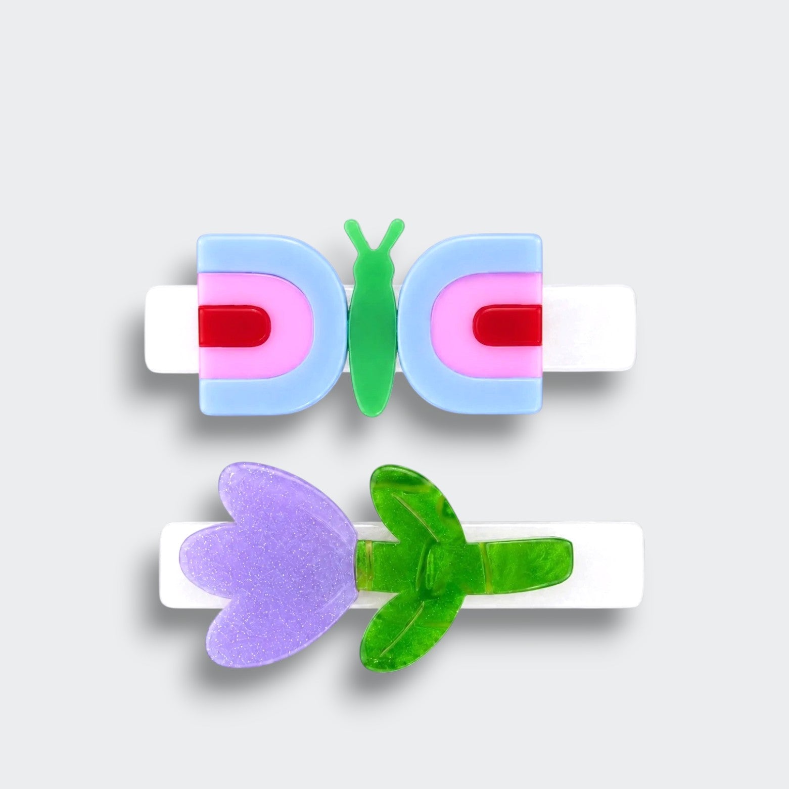 Two white hair clips featuring a blue and pink butterfly and a glittery purple tulip with green leaves on a light gray background.