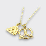 Close-up of a gold-tone pizza slice and crystal-accented pretzel charm necklace on a light gray background.