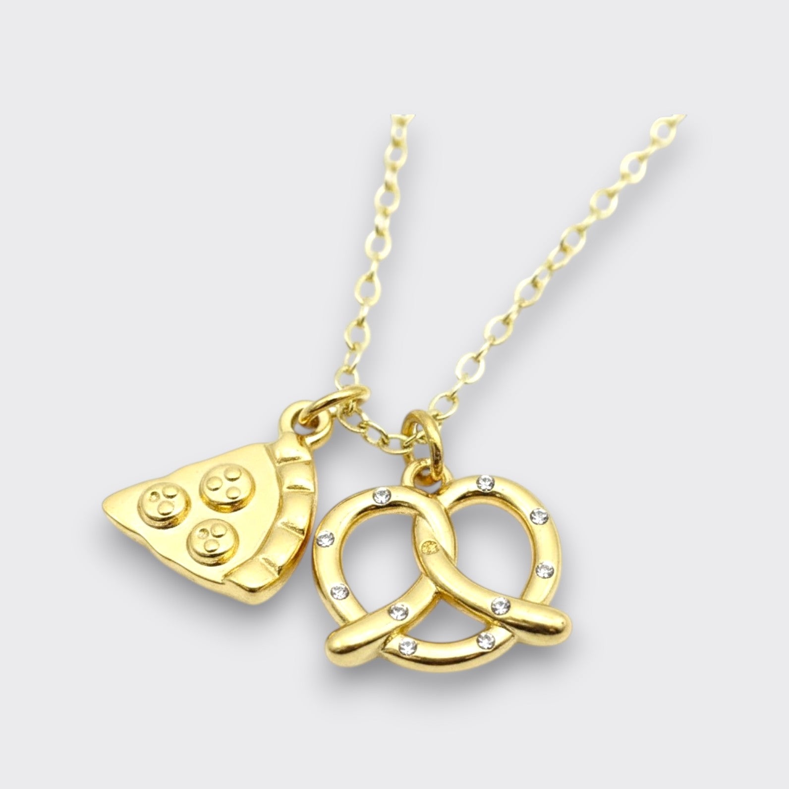 Close-up of a gold-tone pizza slice and crystal-accented pretzel charm necklace on a light gray background.