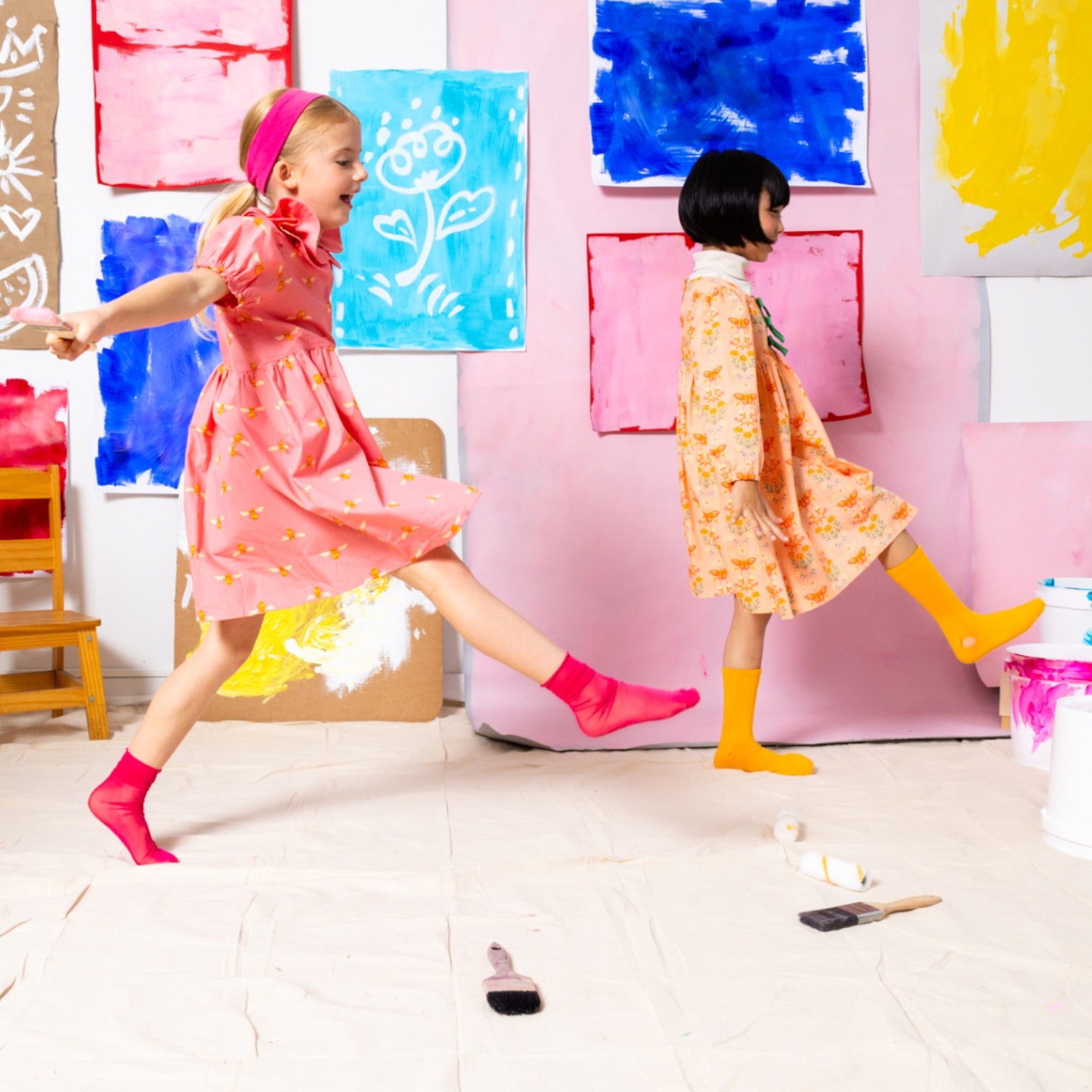 Two young girls wearing Little Moony cotton dresses, one in Honey Bee Petal Double-Collar design and the other in a different playful design, with colorful background, stylish kids’ fashion outfits