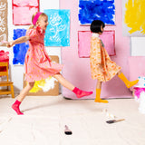 Two young girls wearing Little Moony cotton dresses, one in Honey Bee Petal Double-Collar design and the other in a different playful design, with colorful background, stylish kids’ fashion outfits