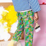 Person wearing colorful floral pants by Little Moony with a striped shirt, holding a paintbrush against a pink background