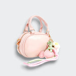 Angled view of a pink bowler purse with a zip closure, heart and floral charms, and trailing pastel ribbons on a light gray background.