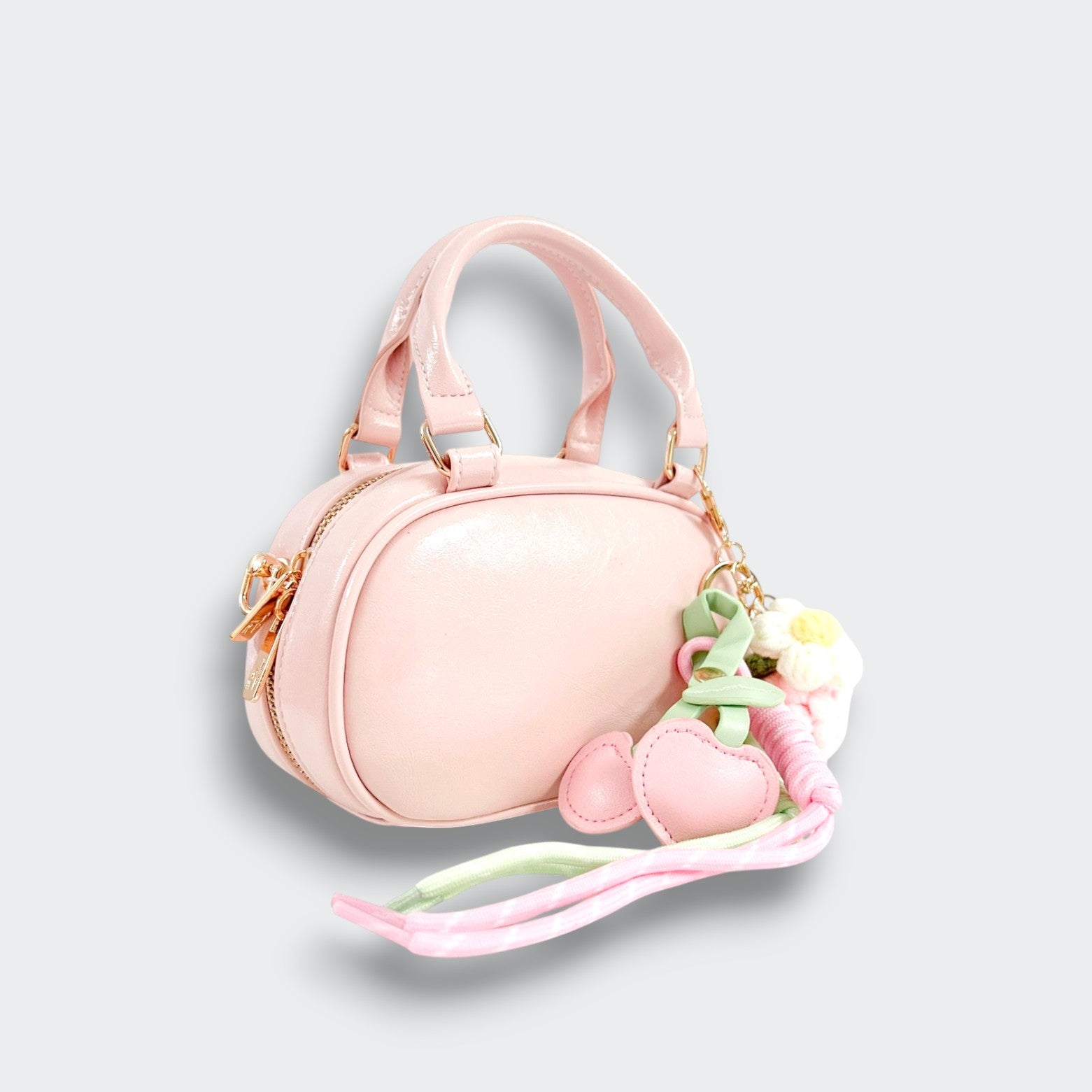 Angled view of a pink bowler purse with a zip closure, heart and floral charms, and trailing pastel ribbons on a light gray background.