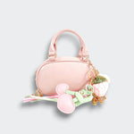 Pink bowler purse with heart and floral charms, pastel ribbons, and a gold-tone chain on a light gray background.